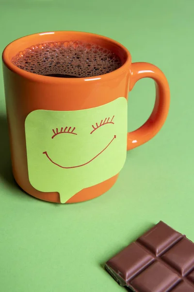 Fresh hot coffee in an orange mug and a funny smiley face sticky note on a green table beside a big piece of chocolate
