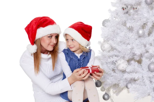 The pretty girl and her mother with  gift box are sitting near Christmas tree on a white background.