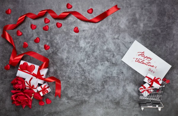 グレーの背景にショッピングトロリー ギフトボックス 装飾的な赤いハート サテンリボンとグリーティングカード コピースペース付きのトップビュー バレンタインデー — ストック写真