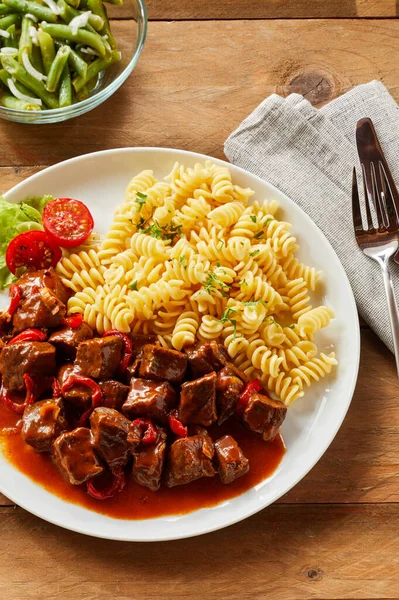 Wild boar or venison stew or goulash in a thick rich gravy seasoned with chili and served with pasta noodles and fresh tomato in an overhead view