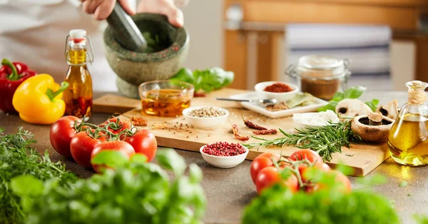 Panorama banner of the hands of a chef preparing herbs by blending them in a pestle and mortar with assorted spices and fresh vegetables on a wooden board