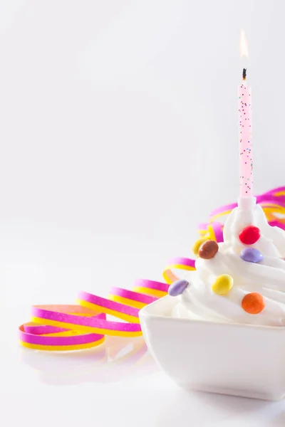 カラフルな丸いキャンディーとコピースペースのピンクのパーティーリボンがトッピングされたソフトクリームのボウルで誕生日キャンドルを燃やす — ストック写真