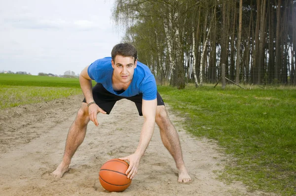 一名身穿蓝色T恤 黑色短裤 弯腰驼背的年轻人的正面全景照片 一只手靠在膝盖上支撑身体 另一只手靠在前排 看着镜头中的篮球 — 图库照片