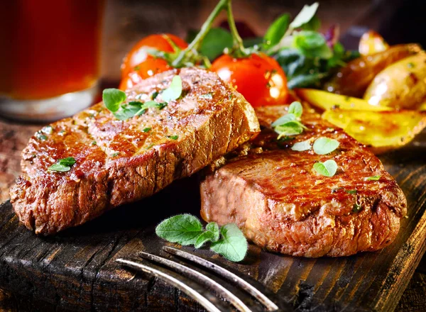 Succulent thick juicy portions of grilled fillet steak served with tomatoes and roast vegetables on an old wooden board