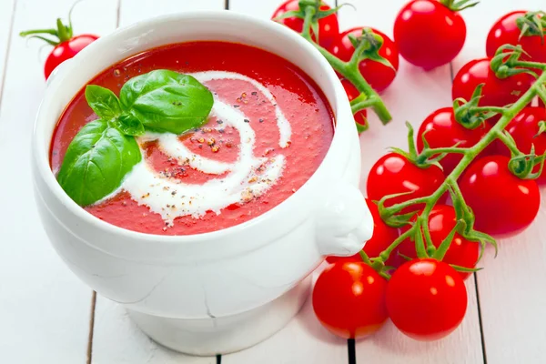 Colourful display of tomato soup with cream and basil alongside a bunch of fresh grape tomatoes on the vine on white painted wooden boards