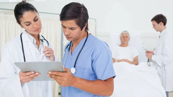 医者と患者の前に立っている間にクリップボードを調べる聴診器付きの医者とインターン — ストック写真