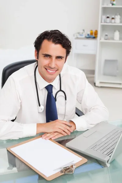 Uśmiechnięty Lekarz Siedzący Przy Biurku Schowkiem Laptopem — Zdjęcie stockowe