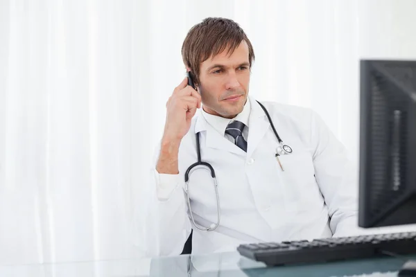 Allvarlig Läkare Tittar Sin Dator Medan Talar Sin Mobiltelefon — Stockfoto