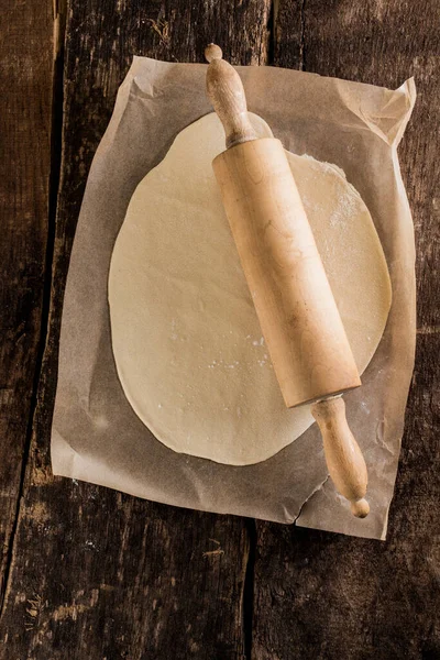 Preparing the base for an Italian pizza with an overhead view of rolled out dough on oven paper with an old-fashioned wooden rolling pin on a rustic wooden kitchen table