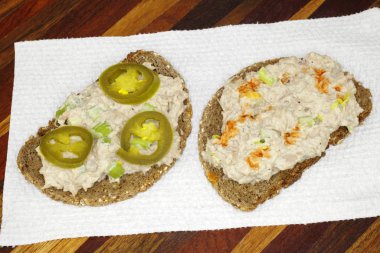 Open faced tuna salad sandwich meal made with celery, mayonnaise, seasonings, jalapeno slices and hot sauce on whole grain pumpernickel rye bread served on a white paper towel on a wood tray. clipart