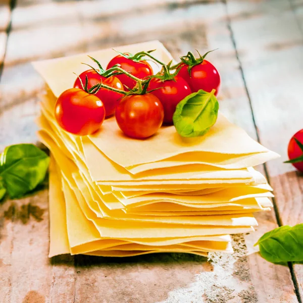 Dried uncooked lasagne pasta sheets with fresh ingredients including cherry tomatoes and basil leaves, close up on old wooden boards