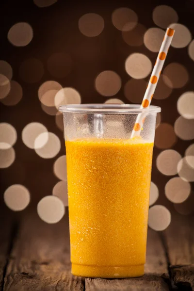 一杯健康而清凉的橙红色芒果冰沙 一根稻草放在木制柜台上 点缀着节日的彩灯 — 图库照片