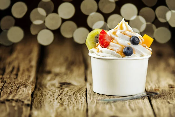 Creamy vanilla frozen yogurt topped with fresh tropical fruit served in a plastic takeaway tub with a disposable teaspoon on a wooden table with a background bokeh of party lights
