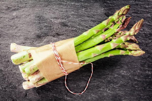 Bundle of farm fresh green asparagus spears tied with brown paper and string on a dark textured slate background, overhead view