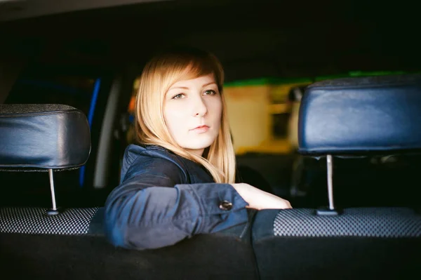 Portrait of a blonde woman sitting in a car in the back seat, placing elbow on the back of the seat, picture taken through the back of the car