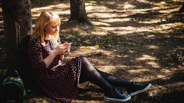 blonde woman dressed summer dress, black pantyhose and sneakers with white sole, resting nature enjoying warm sunny day. Sits under tree alone, holding in hand cell phone, Emotion online communication