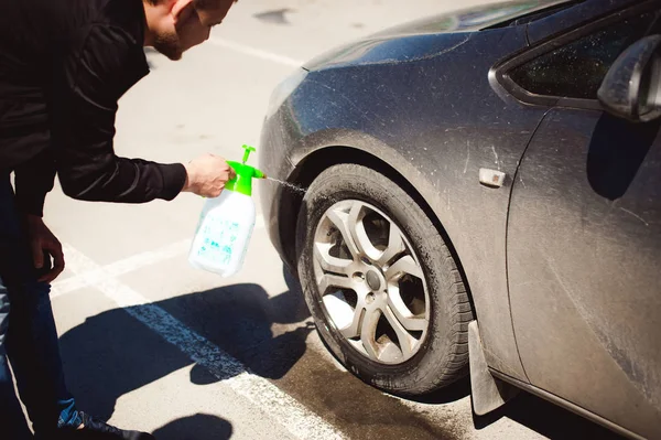 young bearded man washes his car\'s wheel rims, spraying water from spray, in street parking