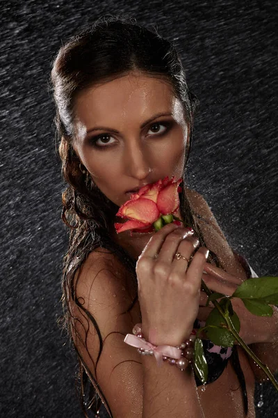 Young beautiful woman with rose stands under rain on a black background.