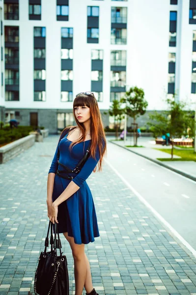 年轻迷人的女人 长长的腿 长长的头发 蓝色典雅的连衣裙 手里拿着黑包的剑带 漫步在欧洲一个现代化的庭院里 一个温暖的夏日 — 图库照片