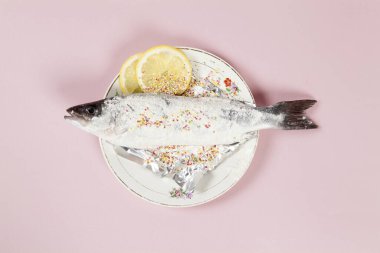 A preparation of a bass fish with sprinkles and a slice of lemon beside inside a flower plate hidden on a pop pink background. Minimal color still life photography. clipart
