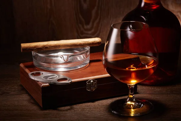 Still life with bottle of cognac, box with cigar and glass with cognac standing on a table on a wooden background.