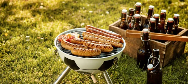 Soccer World Cup themed barbecue with pork and beef sausages grilling over a fire in a soccer ball BBQ and crate of beers in a green field in panorama format