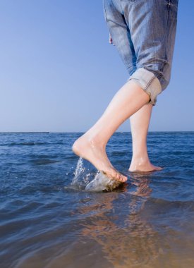 Leg view of a middle-aged woman wearing high-brimx jeans barefoot on the beach in shallow water clipart