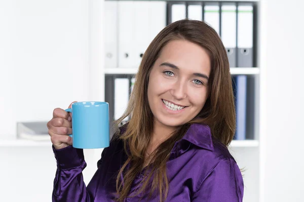A young,smiling woman presents a blue cup while standing in the office. In the background there is a shelf. The woman looks at the camera.