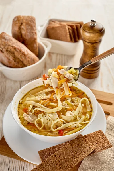 Noodle soup. Delicious soup with noodle, pieces of chicken and vegetables in white bowl served with spices and breads on light wooden background.