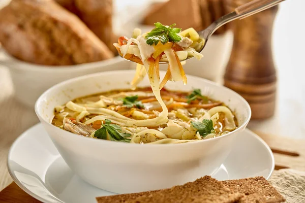 Noodle soup. Delicious soup with noodle, pieces of chicken and vegetables in white bowl served with spices and breads on light wooden background.