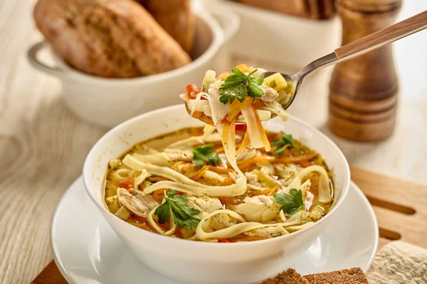 Noodle soup. Delicious soup with noodle, pieces of chicken and vegetables in white bowl served with spices and breads on light wooden background.