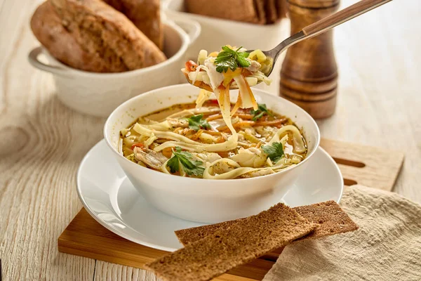 Noodle soup. Delicious soup with noodle, pieces of chicken and vegetables in white bowl served with spices and breads on light wooden background.