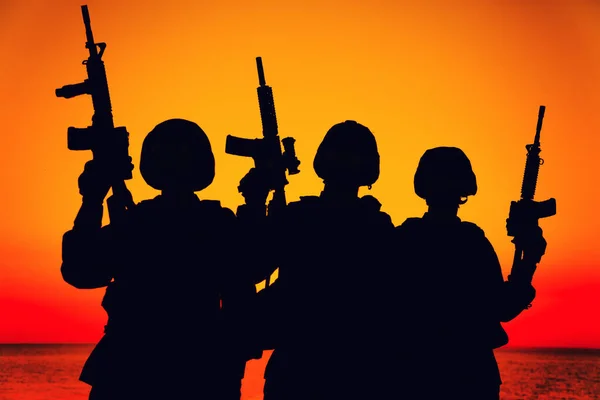 軍隊のシルエット特殊作戦部隊の兵士チーム 海兵隊または沿岸警備隊の戦闘機のグループは 弾薬の乗組員と上昇したアサルトライフルで日没時に海岸に立ってヘルメットを戦う — ストック写真