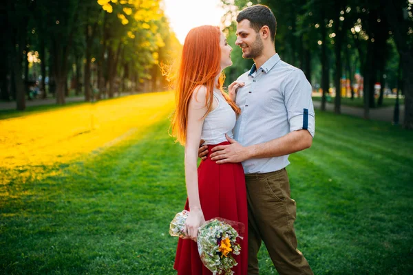 美丽的恋人在日落时拥抱 在户外浪漫地相遇 在公园里 一个带着鲜花的迷人女人和一个年轻男人在一起休闲 — 图库照片