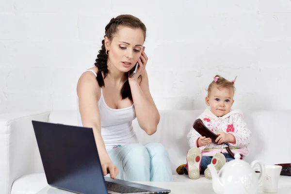 小さな子供とノートと電話を持つ忙しい若い女性が部屋のソファに座っています 母親と赤ん坊 隔離期間隔離社会的距離 リモートワーク — ストック写真