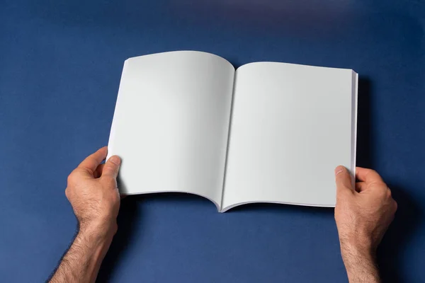 青い背景に空白のページを持つ開かれたブックカタログを保持する男性の手 あなたのデザインのために準備ができてモックアップシリーズテンプレート ページ選択パスが含まれています — ストック写真