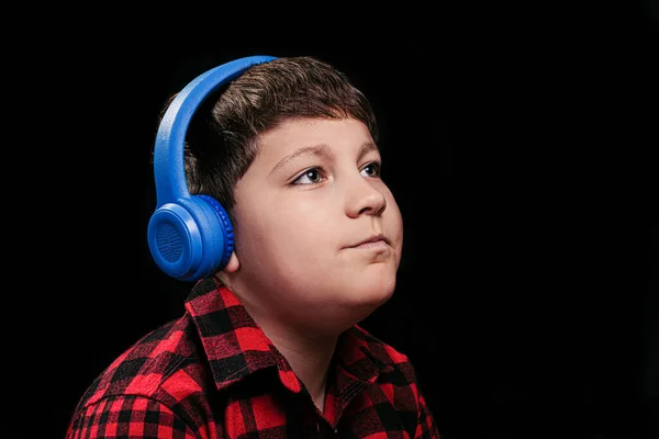白人男の子ティーンでスタイリッシュなプレイシャツで赤で音楽を聴くで大きな青い無線耳のヘッドフォンで隔離された黒の背景 — ストック写真