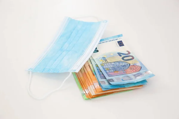 白いテーブルの上に青い外科マスクの下に横たわるユーロ銀行券のヒープ 医療用コロナウイルスのパンデミックの経済的影響の概念 疫学的措置の費用 — ストック写真
