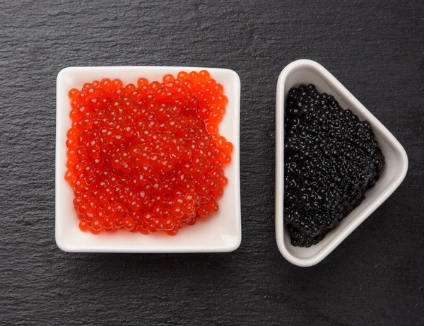 grainy caviar of paddlefish fish in a white ceramic plate and red chum salmon caviar in a white ceramic bowl, black background, top view