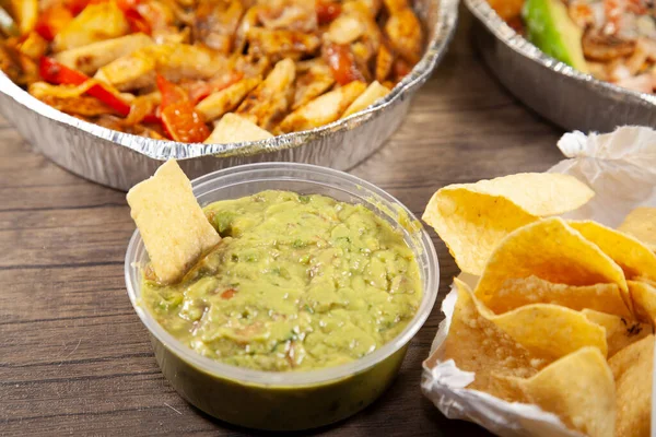 Tortilla chip in guacamole next to an open bag of chips and two takeout meals in tin