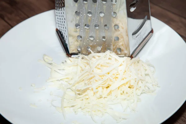 Pile of shredded white cheese on a white plate next to a large, metal cheese grater