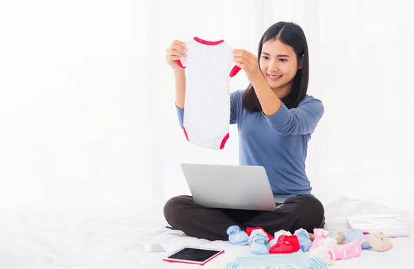 アジアの母ショッピングオンラインのために彼女の赤ちゃん服上のラップトップコンピュータとタブレット上のベッド彼女は新しい子供のための準備のための生まれていない赤ちゃんのための新しい赤ちゃん服を購入します — ストック写真
