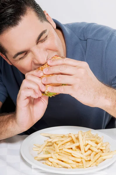 フライドポテトと不健康な食べ物でバーガーを食べる男 — ストック写真