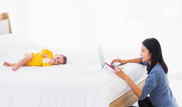 アジアの若い女性ありますワーキング忙しいです上のラップトップコンピュータとタブレットで自宅オフィスでベッドルームで彼女の小さな赤ちゃんあります眠っている間に彼女の小さな赤ちゃんが横たわっていますザホワイトベッド近くのお母さん — ストック写真