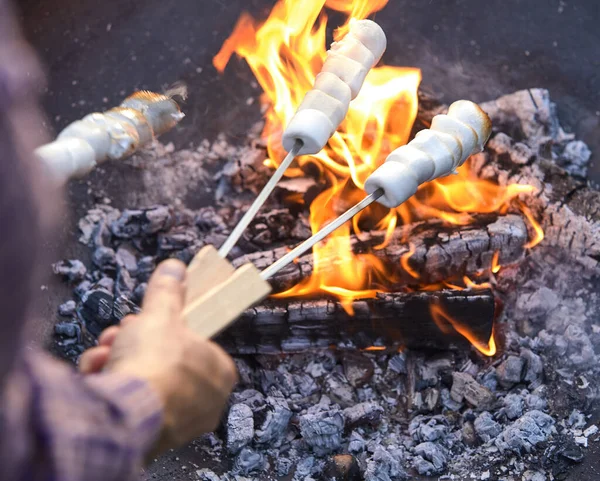 男人在烤肉或篝火的炽热的煤块上 用金属棒烤着发烫的棉花糖 好让糖果从他的肩上掠过 — 图库照片