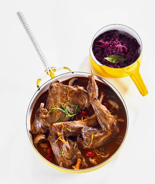 Leg portions of wild rabbit venison in a savory sauce seasoned with orange zest and herbs viewed top down on white with a pot of red cabbage as sides