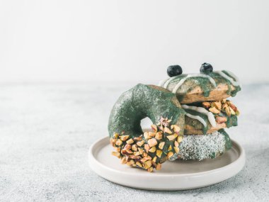 Vegan doughnuts with chia seeds topped with healthy spirulina glaze with pistachio, desiccated coconut and blueberry. Stack of blue green spirulina donuts on gray background. Copy space. clipart