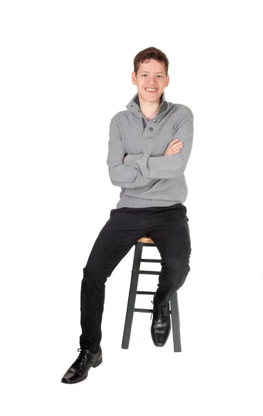 一个年轻的少年 身穿一件灰色毛衣 双臂交叉地笑着坐在椅子上 被白色的背景隔开了 — 图库照片