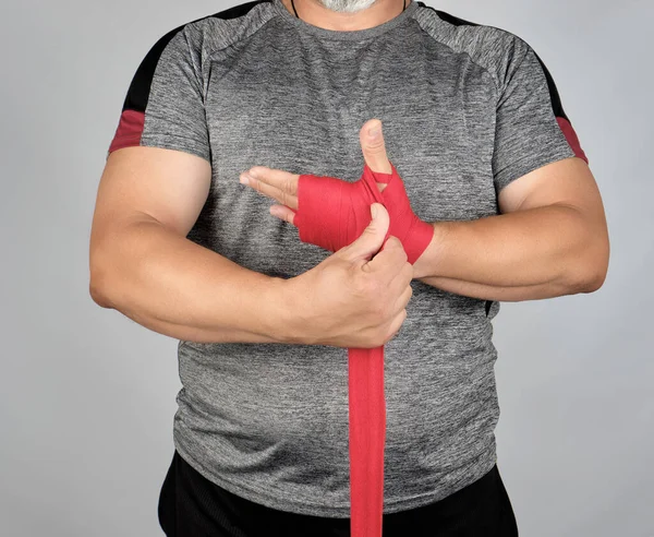 アスリートは灰色の服を着て立ち トレーニング前に赤いテキスタイル弾性包帯で手を包み 灰色の背景 — ストック写真