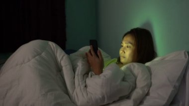 Asyalı kadın gece yatakta akıllı telefonla oyun oynuyor.